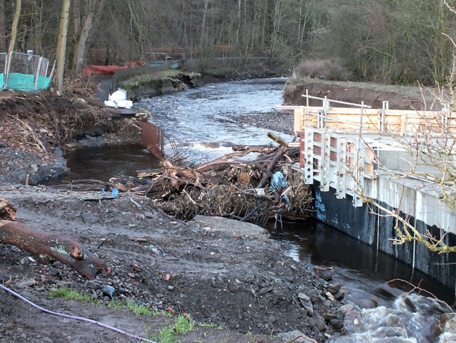 The erosion caused by the blockage can be seen on both banks where the flow diverted around the structure. The steelwork reinforcement can be seen cover with tree trunks and branches and may require some straightening before the work can continue. Upstream, damage to the wall on the Dean Castle bank can be seen in this photo